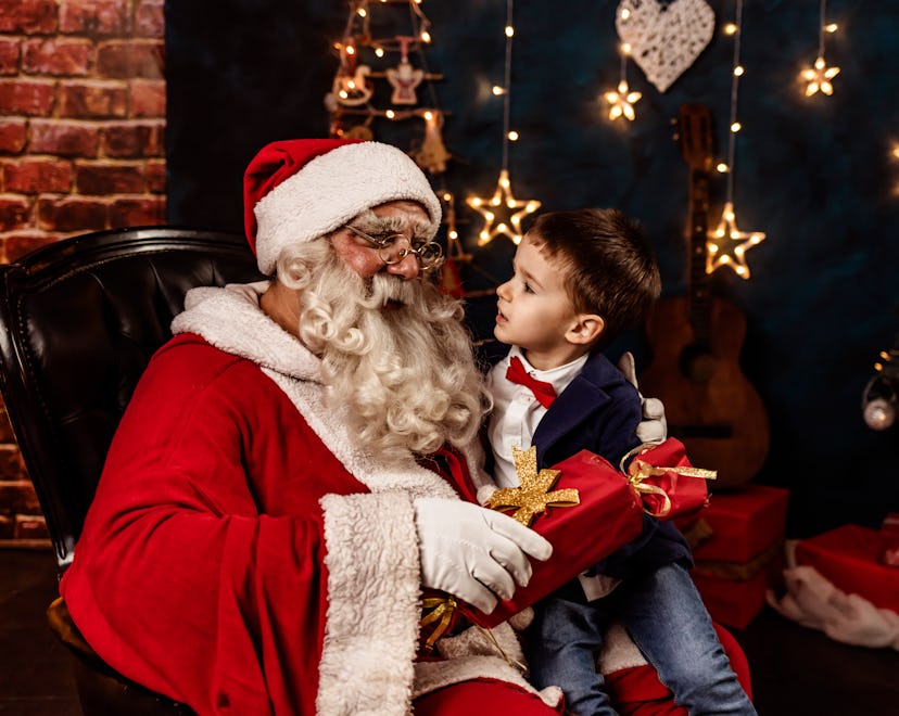 Photo of  little boy sitting in Santa's lap at home and talking to him while holding Christmas prese...