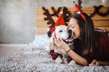 Beautiful young woman sitting on bed with her dog, both wearing reindeer antlers, for which she'll n...