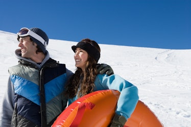 A happy couple smiles while looking off into the distance and holding a snow tube on a snowy hill.
