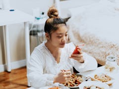 A young woman eats take out food in her bedroom, while posting an Instagram picture on her phone.