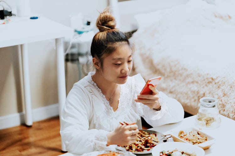 A young woman eats take out food in her bedroom, while posting an Instagram picture on her phone.