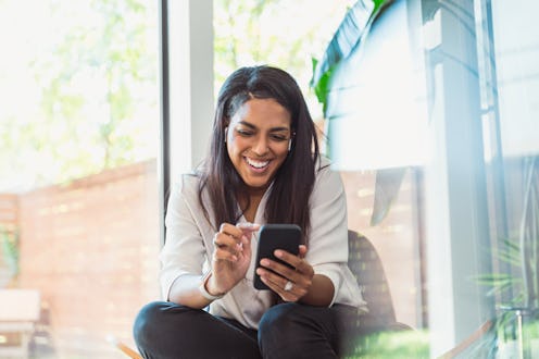A young Black women laughing at a phone, sending a funny text to ask someone out.
