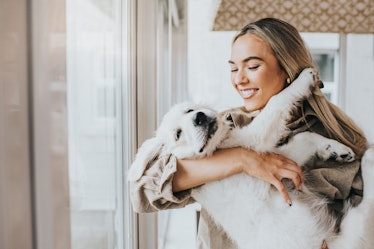 A happy woman holds her dog in a bright living room.