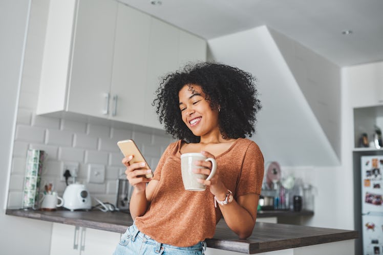 A young woman looks at coffee hacks on TikTok on her phone while drinking coffee in her minimalistic...