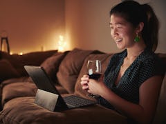 A woman enjoys a glass of wine while sitting on her couch with her tablet in her lap. 