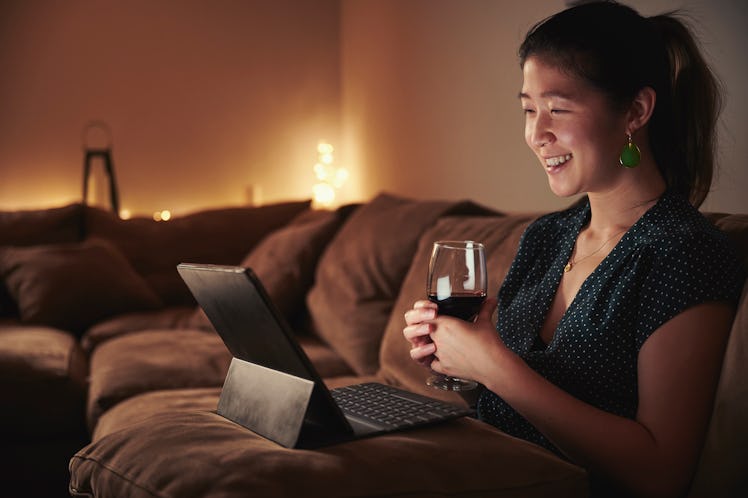 A woman enjoys a glass of wine while sitting on her couch with her tablet in her lap.