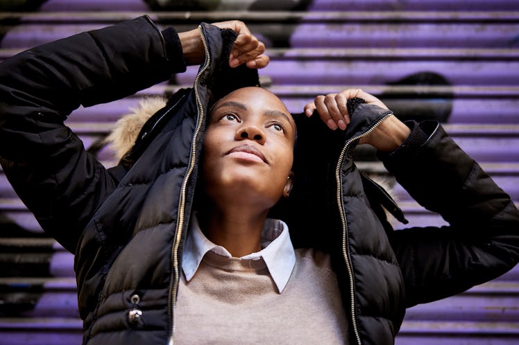 A young woman pulls a black puffer jacket over her head, while posing in front of a purple wall.