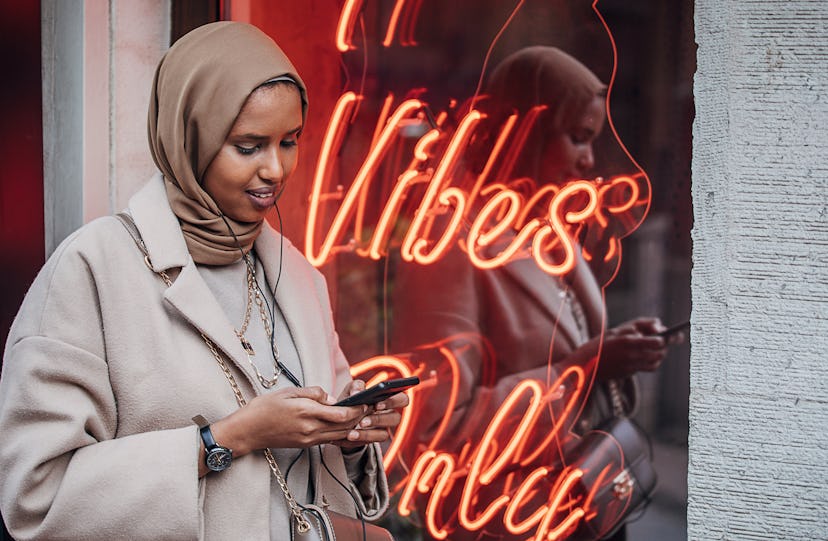A woman wearing hijab checks her messaging apps in front of a neon sign.