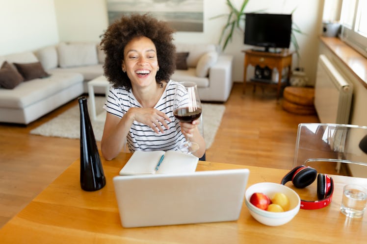 A happy woman laughs while enjoying a glass of red wine and taking a virtual wine tasting class for ...