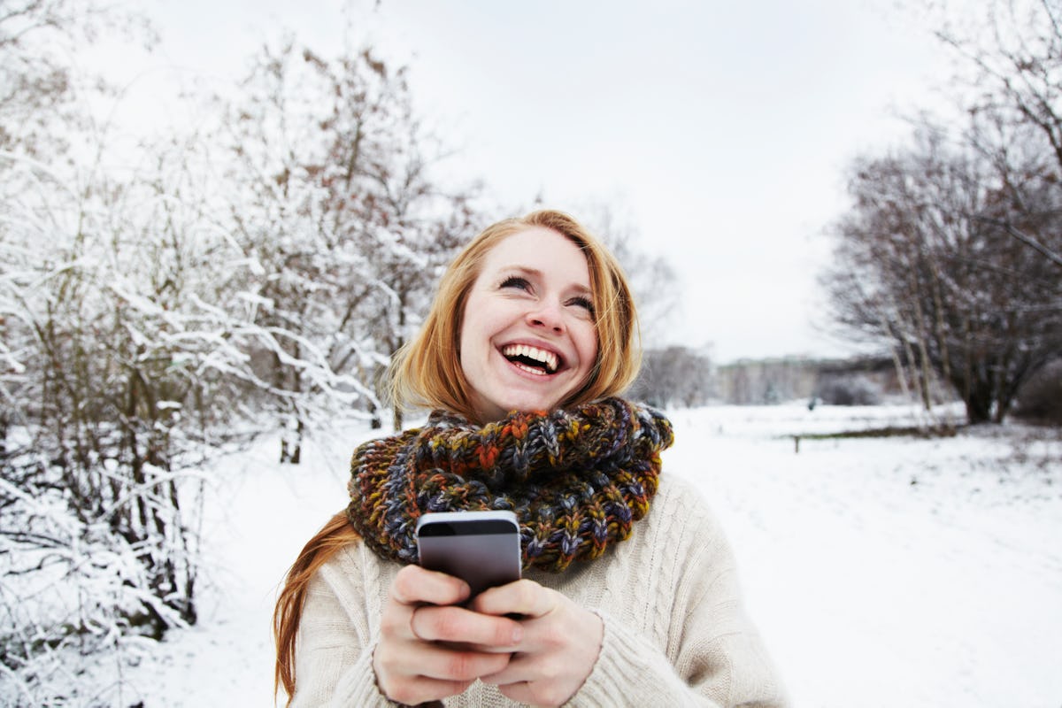 A happy woman laughs while typing on her phone and standing outside in the snow.