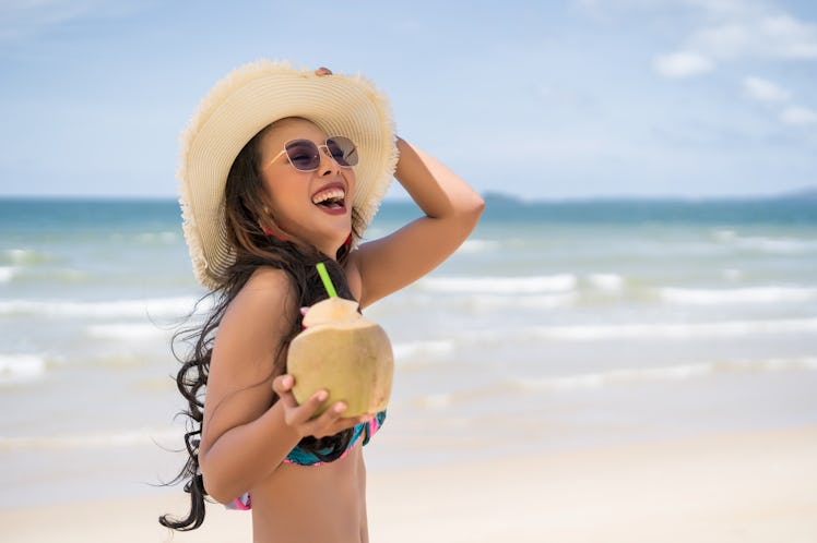A happy woman with a coconut drink walks on the beach with her sunhat on spring break.