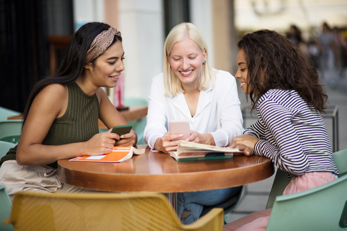 Three friends smile while relaxing at a cafe and looking at one of their phones.