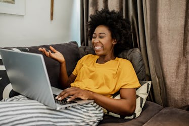 A happy woman dressed in a yellow shirt and striped pants laughs while on a virtual call with her lo...