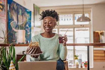 A happy woman holds up a birthday cake and cocktail while on a virtual birthday celebration call.