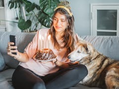 A happy woman holds a birthday cake and poses on the couch with her dog for a selfie.