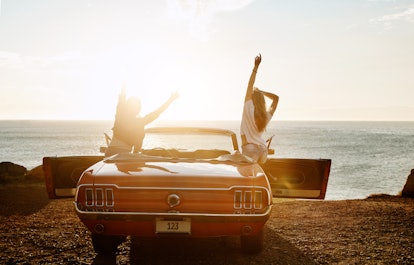 Two girls pose in a red classic mustang by the beach on a road trip.