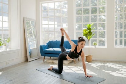 A brunette woman strikes a yoga pose on a mat in her bright home while doing a virtual yoga class.