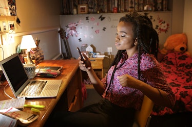 A young woman looks at her phone while sitting at her desk and completing homework at night.