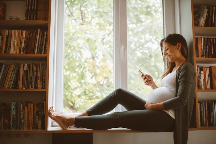 pregnant woman looking at her phone sitting in front of window