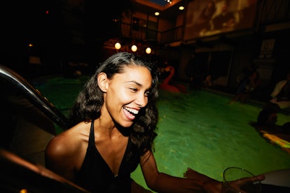 A young woman laughs while standing in a pool at night, in a throwback bathing suit picture.