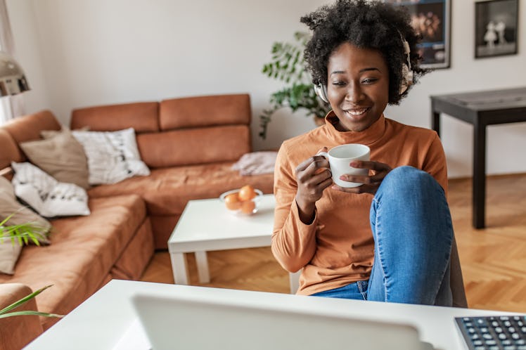 A cozy woman sips from a mug and chats on her laptop while at home.