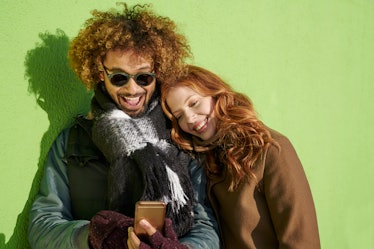 A happy couple dressed for winter leans against a lime green wall and smiles while texting in a coup...