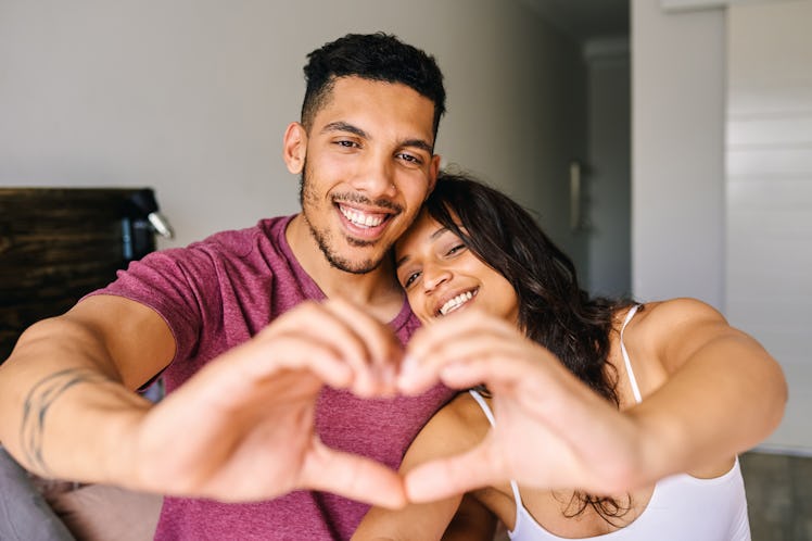 A happy couple in loungewear makes a heart with their hands while relaxing at home on Valentine's Da...