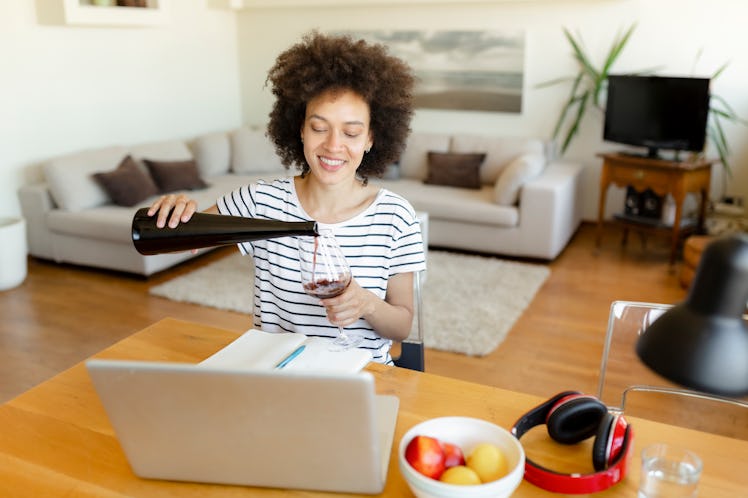 A smiling woman pours wine into her glass in front of her laptop while on a virtual Galentine's Day ...