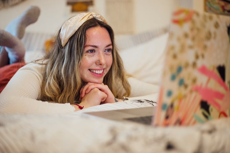 A cozy woman lays in bed in loungewear and smiles at her laptop while celebrating virtual Galentine'...
