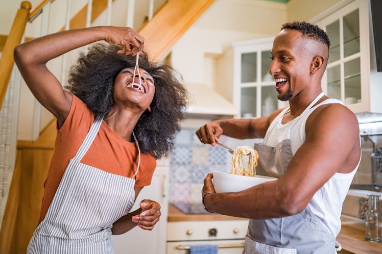 A couple goofs around in the kitchen and eats noodles while making pasta.