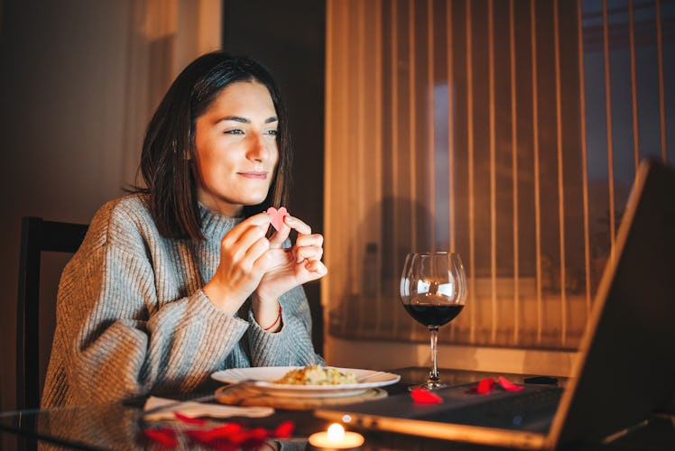 A cozy woman holds a paper heart to her laptop in front of wine and dinner while on a virtual Valent...