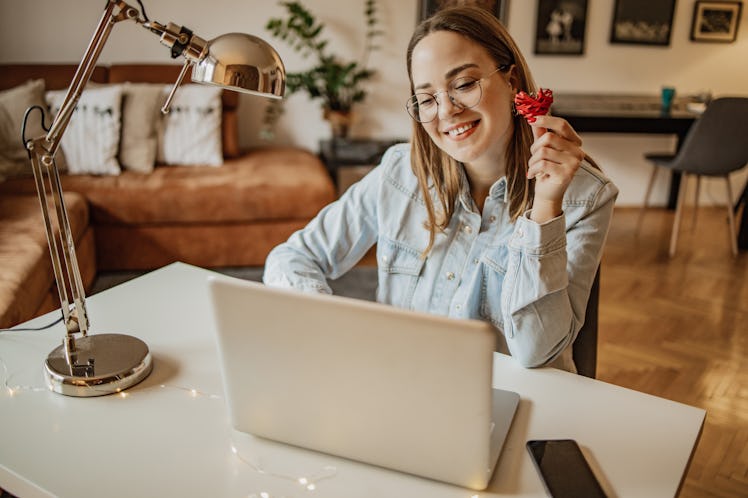 A woman gives a soft smile to her laptop, as she holds up a DIY heart while on a virtual Valentine's...