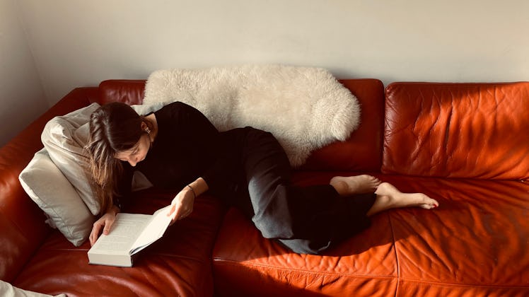 A single woman relaxes on a red leather couch and reads a romance book on Valentine's Day.