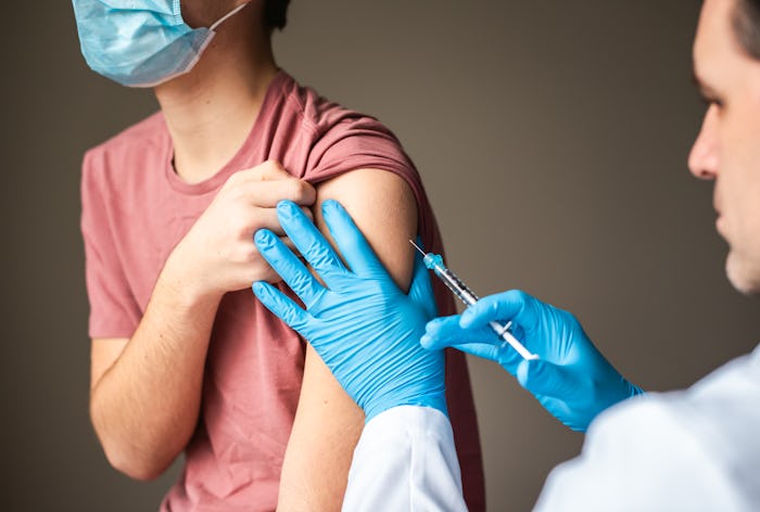A teenager wearing a surgical mask holds up their shirt sleeve as a medical professional wearing blu...