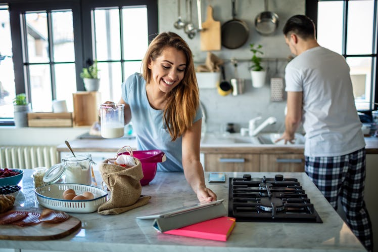 A photo of happy couple preparing breakfast in their kitchen with the young woman looking for a reci...