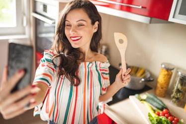 Beautiful young woman using her smart phone and cooking in her kitchen
