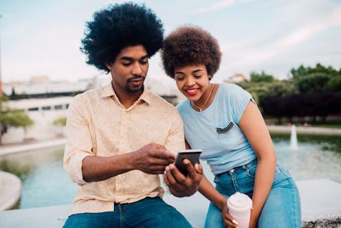 Young African - American couple on vacation sitting in the park and using a smartphone.