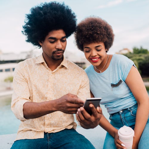 Young African - American couple on vacation sitting in the park and using a smartphone.