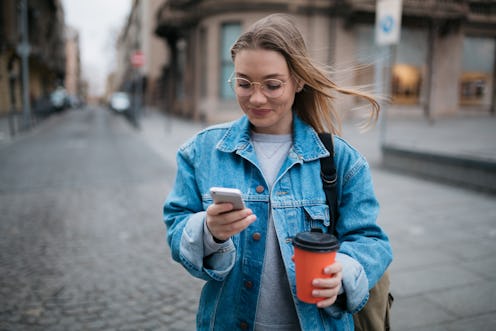 Happy young Caucasian woman using a smart phone outdoors and enjoying a cup of coffee