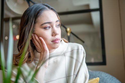 A young woman appears sad and is looking outside her bright window.