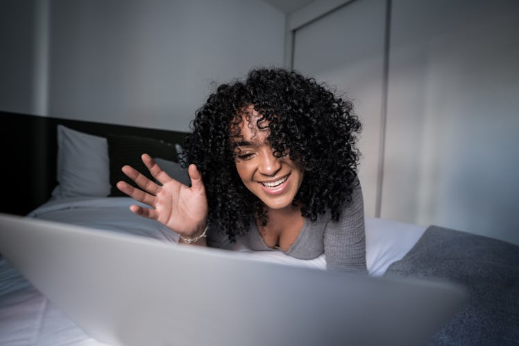 Young woman waving while doing a video call on laptop lying in bed at home