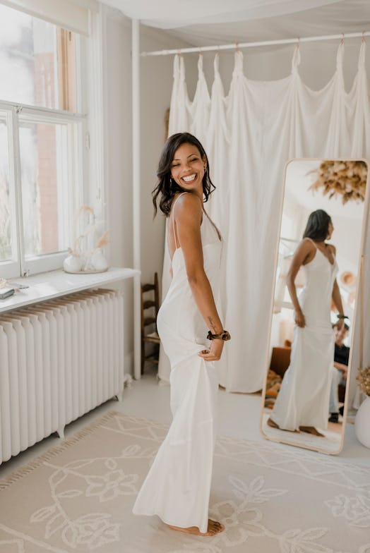 A bride smiles after saying yes to the dress, and standing in the view of a full-length mirror.