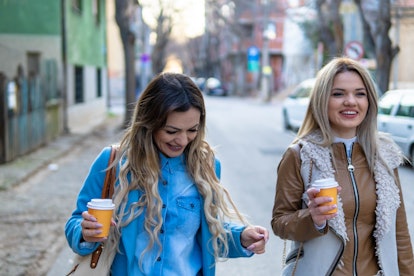 Two young stylish woman, sisters walking by street with coffee cup. Smiling sisters walking outdoors