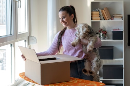 A young woman opens a dog subscription box while holding her fluffy dog.