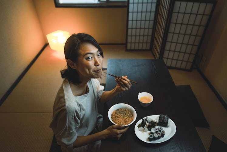 A young woman eats ramen and sushi while sitting at a low table in her home.