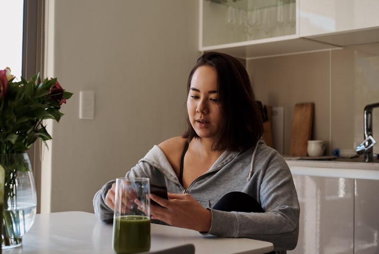 Shot of a young woman using her cellphone and drinking green juice while sitting at home