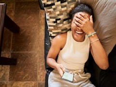 A woman laughing at a text while laying on her couch. 