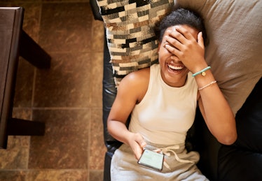 A woman laughing at a text while laying on her couch. 
