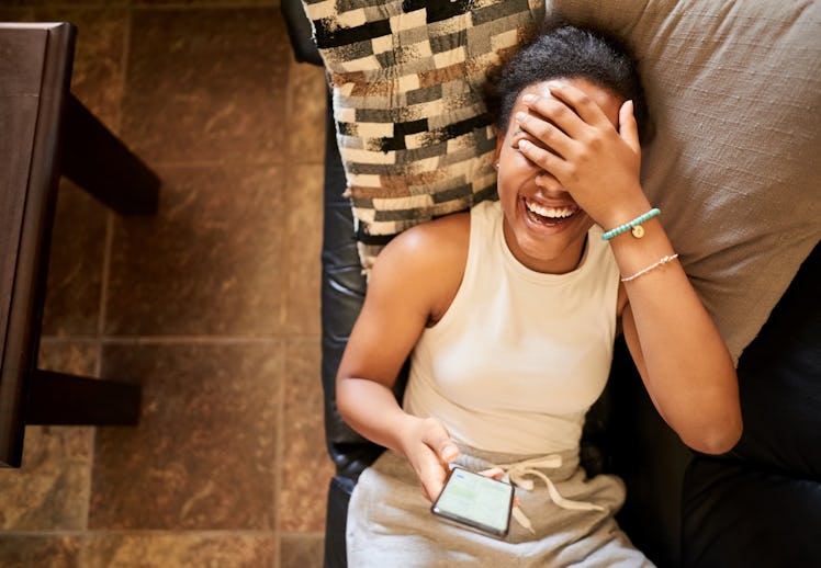 A woman laughing at a text while laying on her couch.