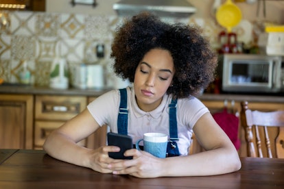 Depressed young afro woman texting on phone alone, sitting sad in the kitchen in her home